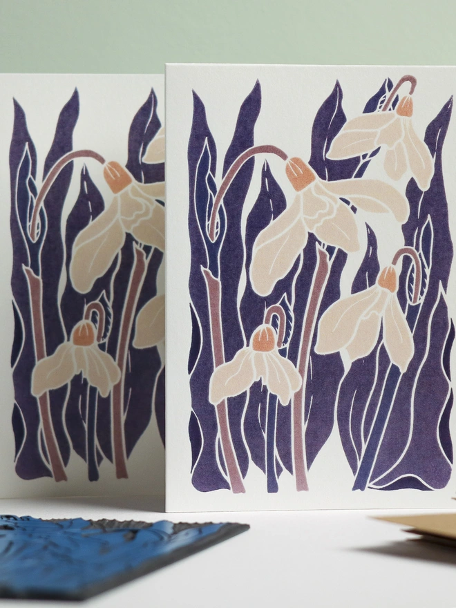 Hand-carved lino block displayed with finished Snowdrops notecard from set of 8. Traditional relief printmaking block showing carved botanical design alongside contemporary printed winter flower cards. Authentic linocut process, Norfolk countryside studio