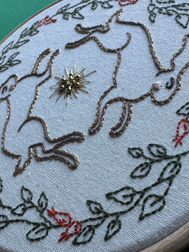 Two hares with pink cheeks and edged in gold jumping a gold bead circled by leaves and rose hips edged in gold 