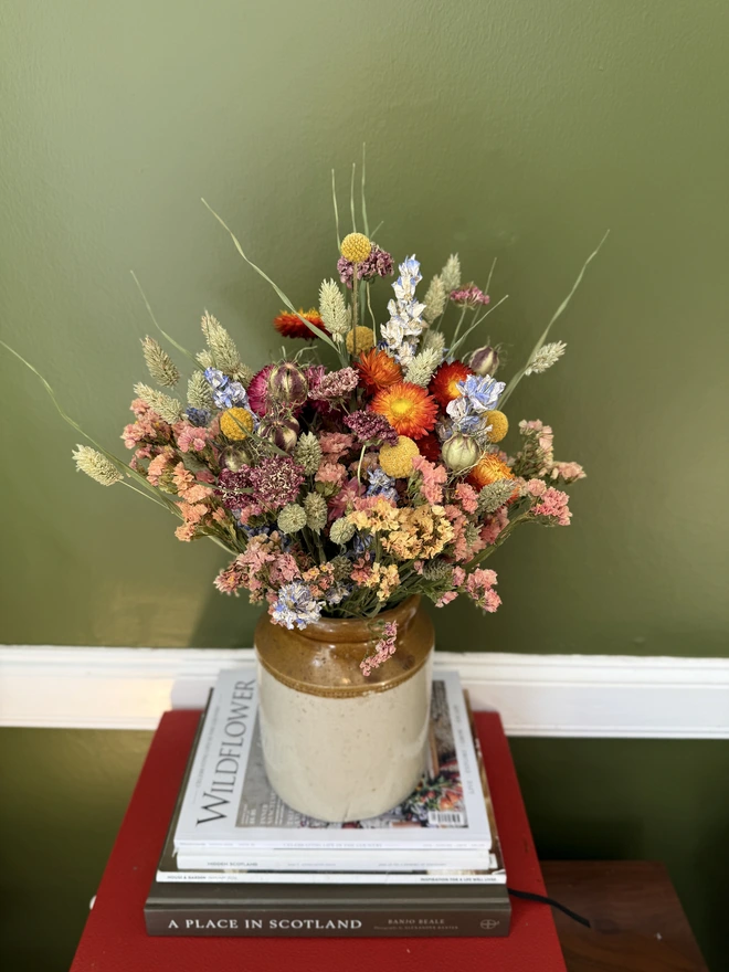 a dried flower bouquet of spring/summer colours in a victorian jam pot sitting on top of a stack of books and magazines against a green wall