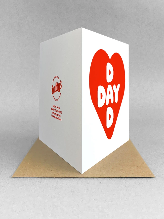 Rear view of a red screenprinted heart card containing intersecting words Dad and Day, a square card stood on a kraft envelopei n a white studio backdrop