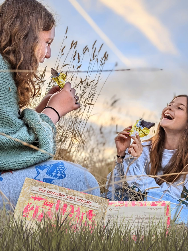 Two happy girls in a field enjoying charity white chocolate bar wrapped in gold foil 