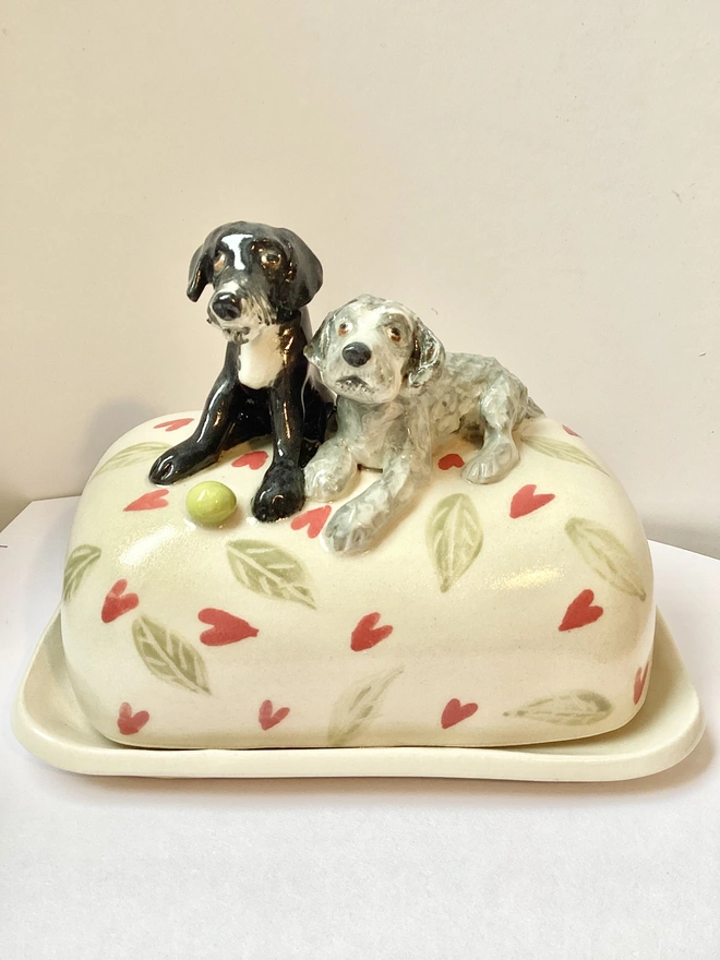 Two dog lying down on a butter dish with a green ball in front of them. Base is cream with red hearts and green leaves. 