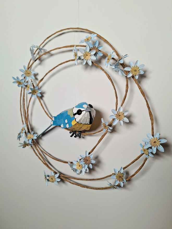 A paper sculpture of a blue tit bird, perched on a floral wreath of blue flowers