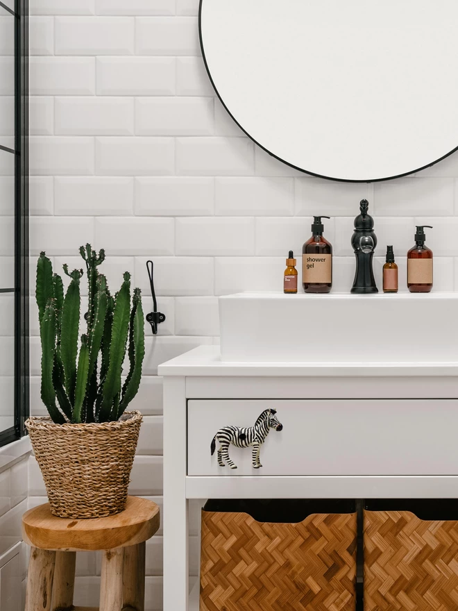 A modern bathroom with white rectangle tiles, a large round mirror with a thin black rim, the edge of a black shower screen and a wide white sink. On the sink vanity unit there is a zebra drawer knob. The brand of zebra animal drawer knobs is Candy Queen Designs. Underneath the zebra drawer knob are two rustic storage baskets and to the left there is a snake plant in a wicker basket sitting on a wooden stool. 