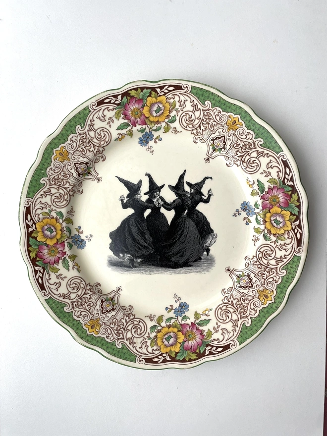a vintage plate with an ornate floral border in the centre is a witch's coven, the four of them are hand in hand dancing in a circle