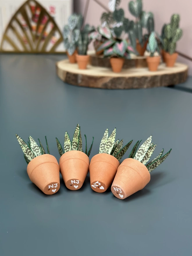 4 miniature replica Sanseveria Snake Plants lying down on a blue desk to show the bottom of the pots which show NJ and a heart written in white
