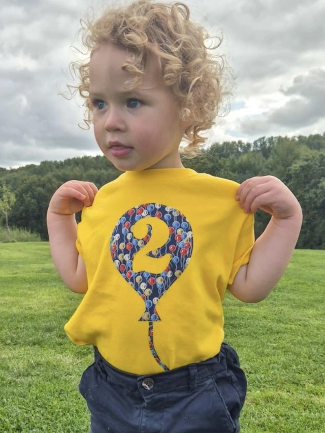 A smiling 2 year old boy wearing a yellow cotton t-shirt. The t-shirt features a balloon with the number 2 cut out from it, appliquéd in a balloon Liberty print.