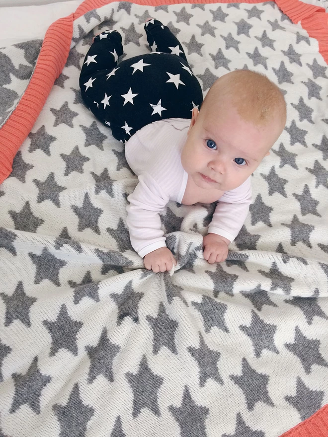 A view of a baby lying on its tummy on a white knitted blanket with a grey star pattern. the blanket has a bright coral pink coloured trim. The baby is looking at the camera and scrunching the soft blanket between her fingers.