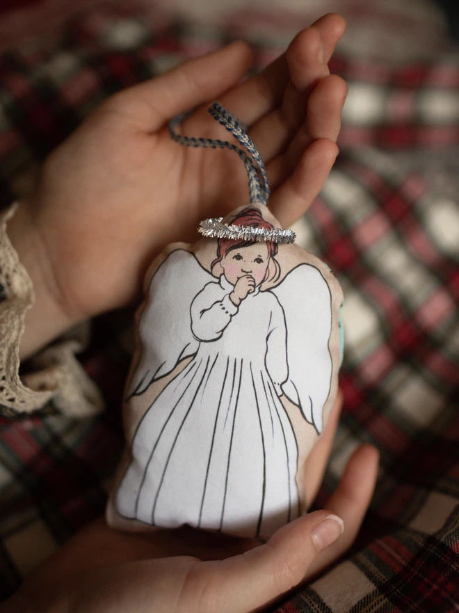 a Childs hands holding an orange haired angel decoration a little girl angel sucking her thumb made from plush printed fabric with a tinsel halo and a ribbon for hanging