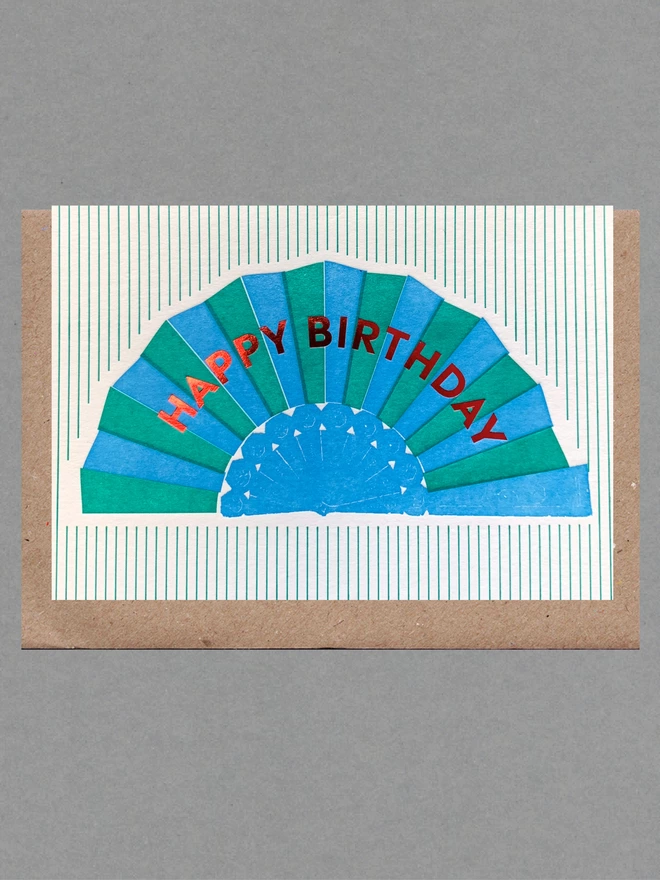 White card with blue and green fan with red text reading 'Happy Birthday' on green striped background with brown envelope behind