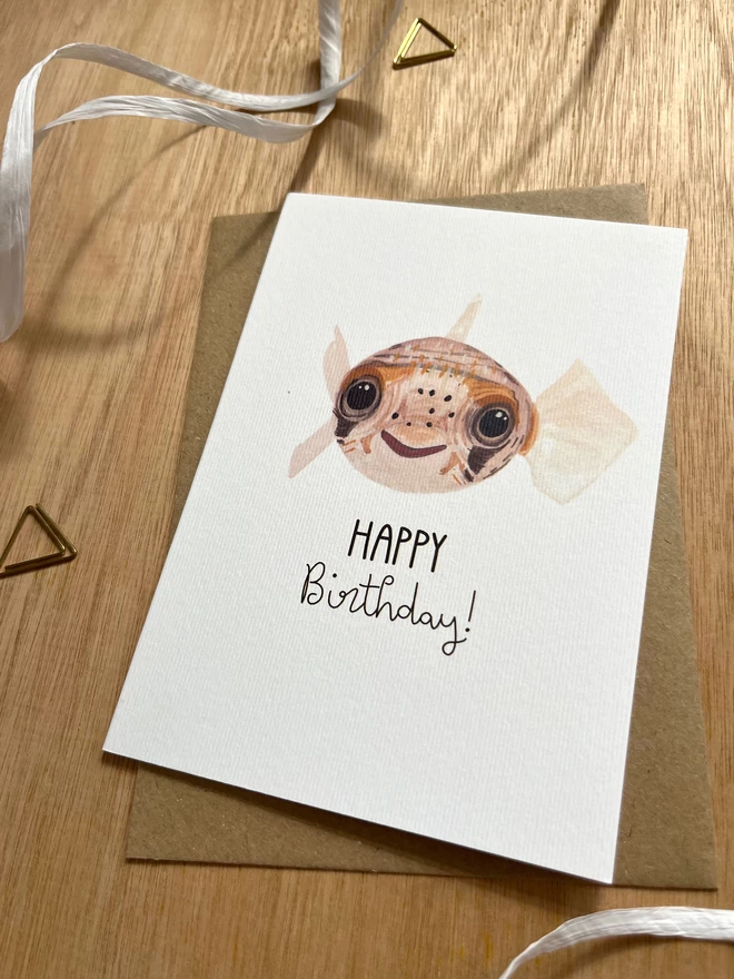 A greetings card with a white background featuring a deflated pufferfish on their back with the phrase "happy birthday" underneath