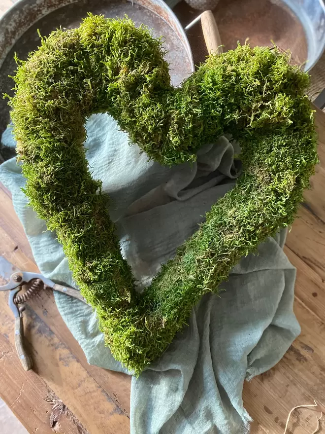 A hand bound moss heart wreath lays against a silver bucket, a duck egg blue cloth drapes over the bucket.. 