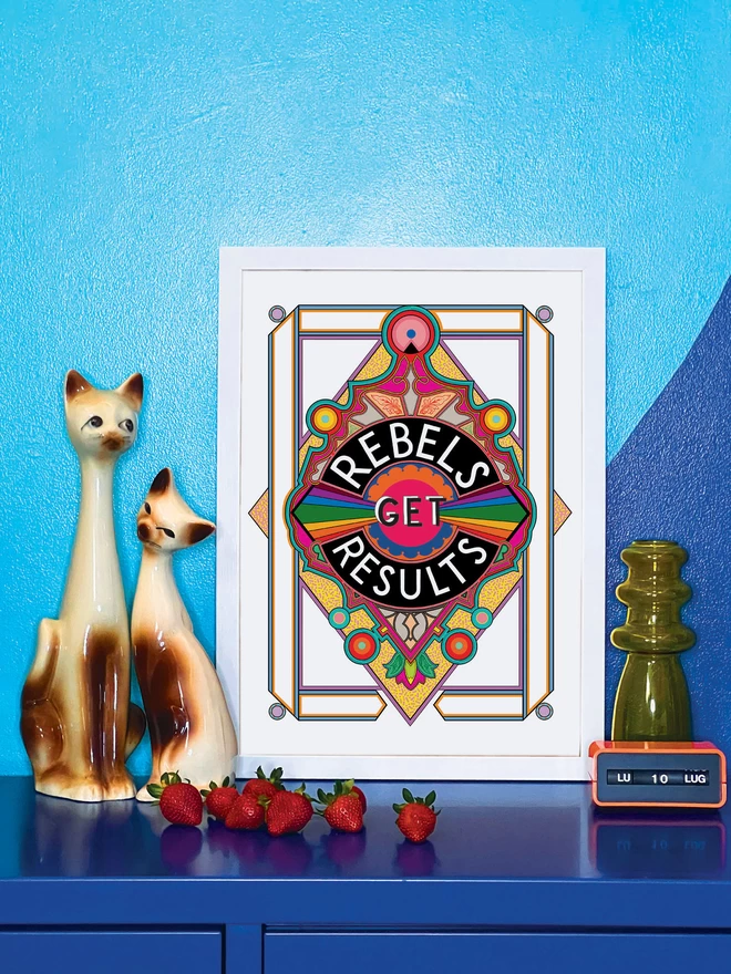 Rebels Get Results is written in white on a black background at the centre of this vibrant, abstract portrait illustration, with a white background and rainbows emitting from the centre, and multi-coloured detailing. The picture is in a white frame, against a turquoise and blue wall resting on a blue cabinet. Next to the picture are two cat ornaments, some ripe strawberries, a yellow glass vase and an orange Italian plastic calendar showing the date as ‘LU 10 LUG’.