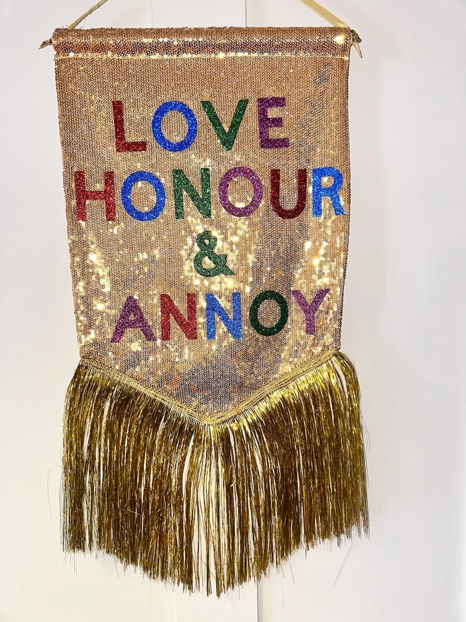 A mini customisable banner hangs from a hook. It has a gold sequin background and a gold tinsel trim along the bottom. The text is multicoloured and says 'LOVE HONOUR & ANNOY'