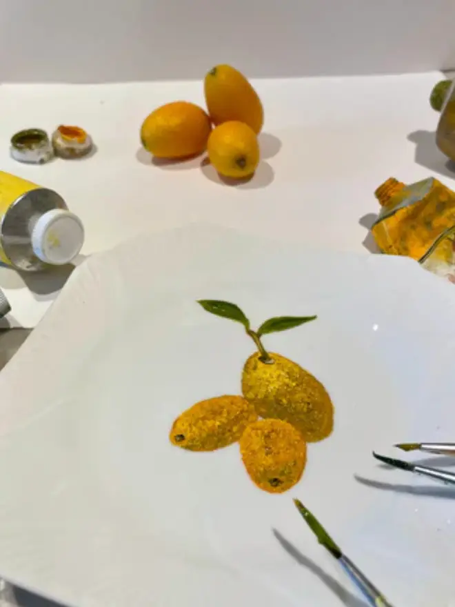 Fine Art Oil Painted Kumquats On A Antique Tea Plate, hanging on a yellow wall, in a studio next to fresh kumquats.