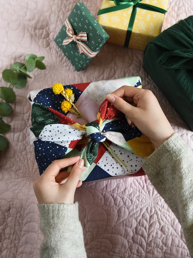 A gift is being wrapped in patchwork cotton fabric wrap, and a small posy of yellow flowers is being tucked into the knot.