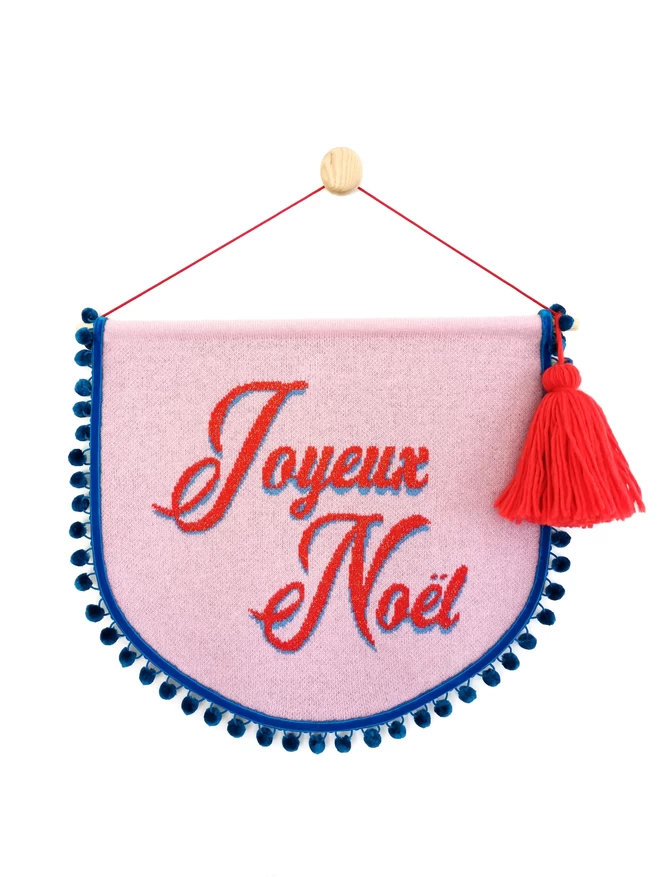 A product image of a pink knitted scallop shaped wall hanging with teal pom pom trim and the words ‘Joyeux Noel’ written across the banner in red retro writing with a hint of gold sparkle in the text.. An oversized red tassel hangs from one side of the wooden doweling and the whole banner is suspended from a wooden wall dot by a bright red nylon cord.