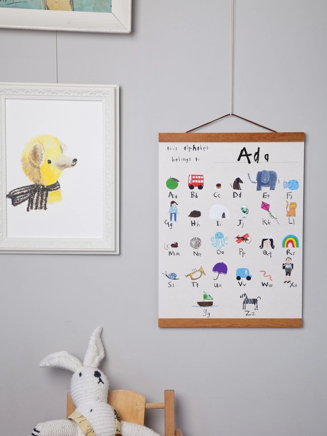 grey wall with alphabet poster hanging on oak poster hanger next to soft toy and framed pictures