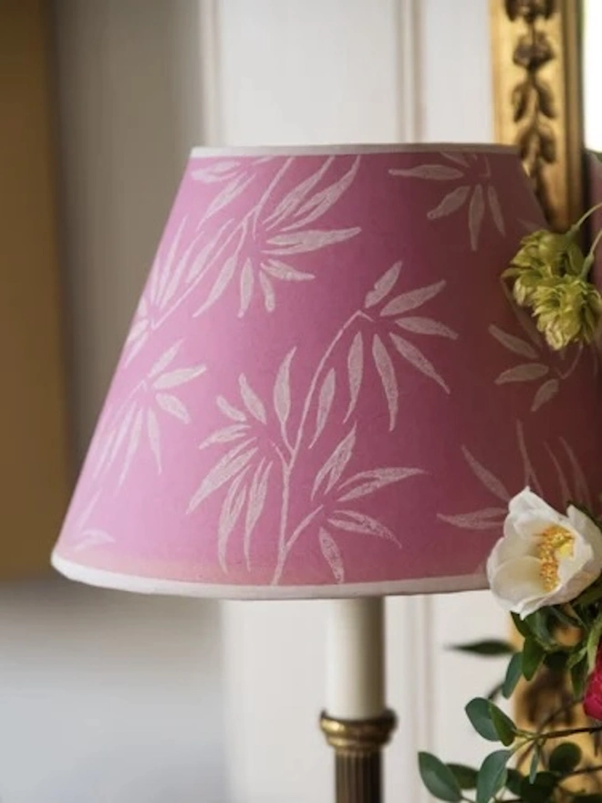 Paper Empire Lampshade. A tapered pink lampshade decorated with a delicate white bamboo leaf print and a pale trim, set beside fresh flowers and a gilt frame. The botanical hand-printed shade adds a soft, romantic touch to a dressing table or living room.