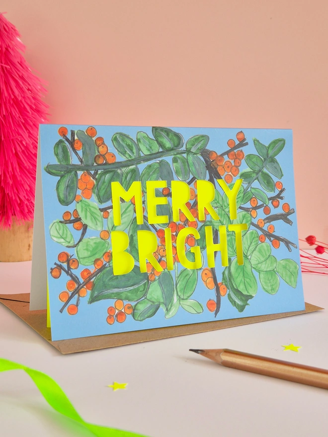 Merry Bright Christmas Card with Orange Berry Illustration, paper cut text with neon yellow slogan that says Merry Bright 