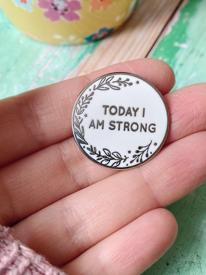 A round white enamel pin with a floral design and the words "Today I Am Strong" rests on a hand above a green desk.