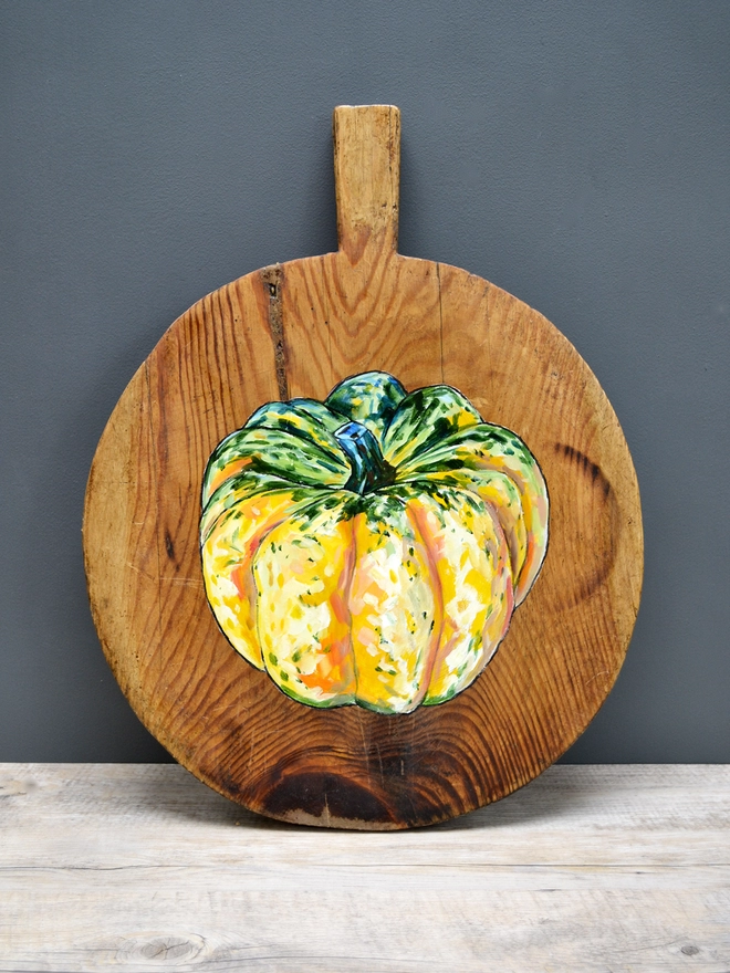 Wooden chopping board with yellow, orange and green squash painted design standing against a wall