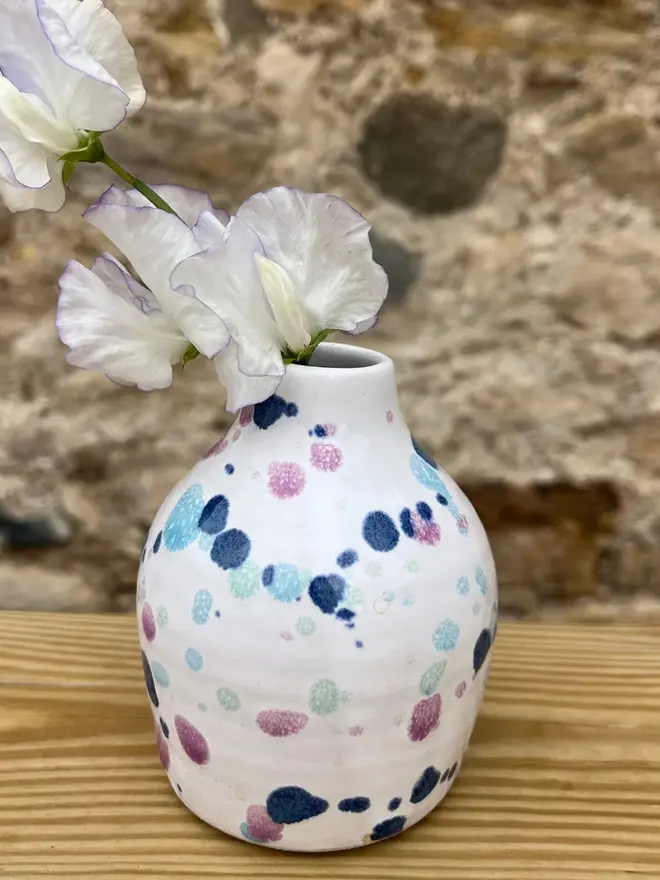 Sweetpea Bud Vase, a colourful vase sitting on a wooden surface against a brick wall backdrop. It is accompanied by a flower. 