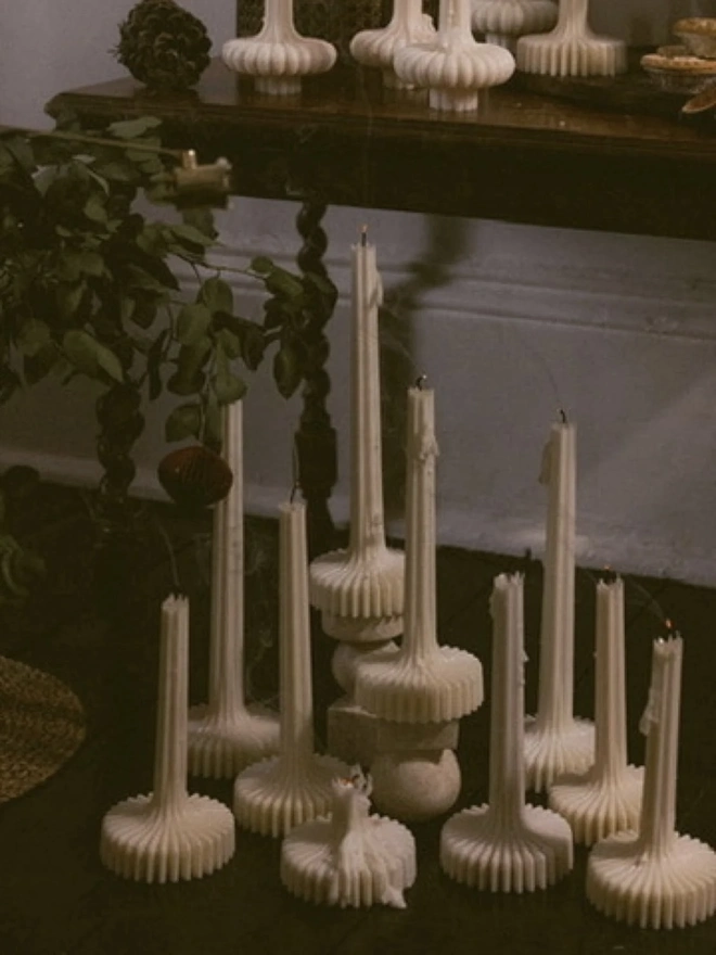 Pair Of White Flair Candles (Set Of 2). A group of cream fluted candles in different heights standing on a dark wooden floor beneath a console table, with eucalyptus and a paper bauble beside them. The flared ribbed bases give a dramatic, architectural look to holiday styling.