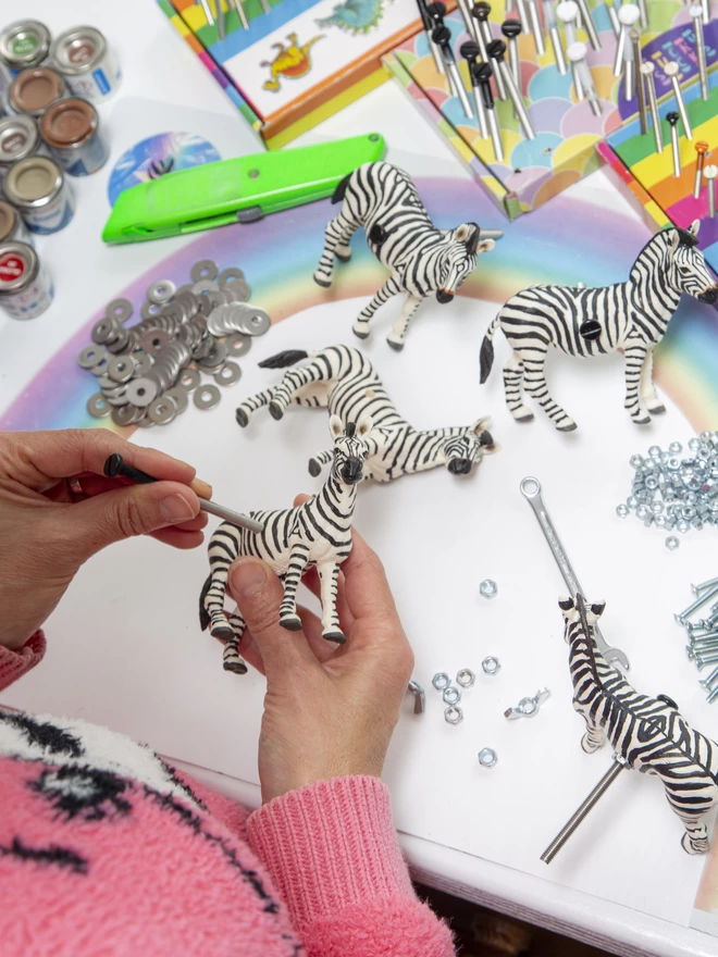 A white work bench with tools and hardware, there are three plastic zebra furniture knobs, a pile of washers and nuts, a small spanner and a stanley knife on it. There are a pair of hands putting the bolt into a zebra furniture knob, there is a pastel rainbow on the worktop.