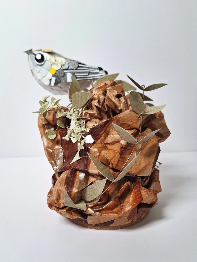 Firecrest paper bird sculpture, perched on a tree stump. Side view.