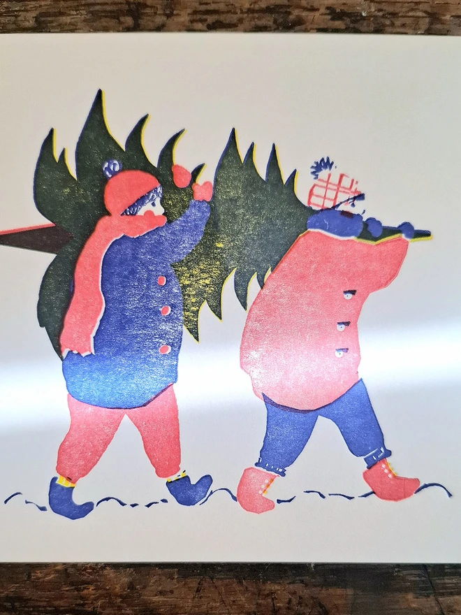 A handprinted greetings card lying flat on a wooden surface, showing the illustration on the cover of two figures carrying a Christmas tree on their shoulders, and printed in red, blue and yellow.