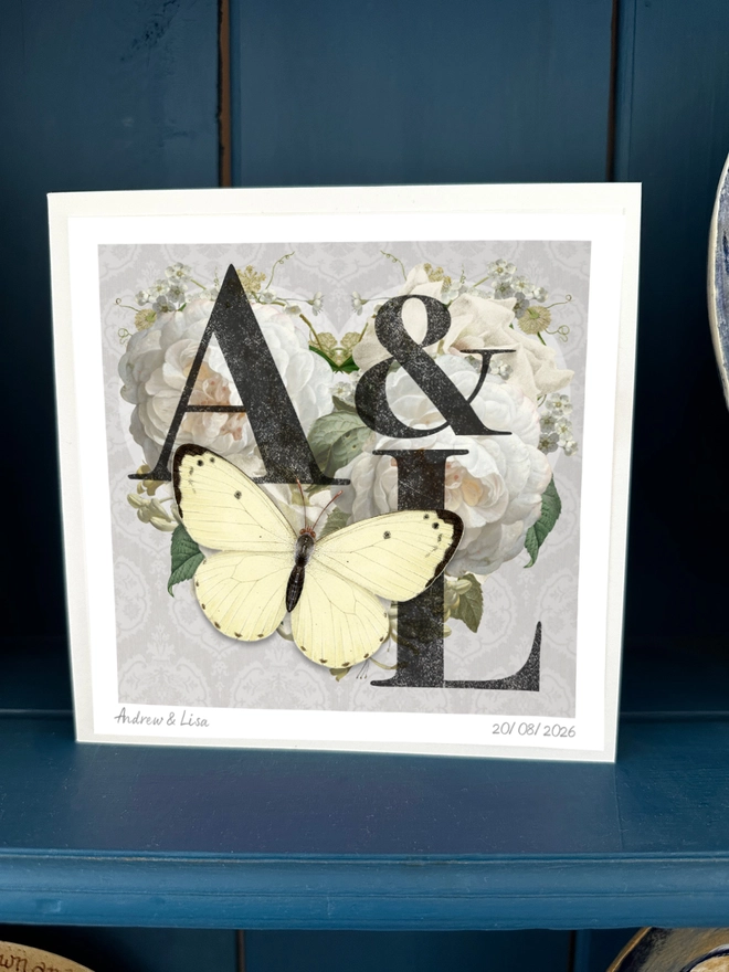 A handmade Diamond Wedding anniversary card featuring two initials of a couples names, place inside a heart design with vintage roses and on a silver grey background. The recipients initials are hand written on the front of card and hand signed, A white 3D papercut butterfly sits on the heart and wings can be folded out for display.