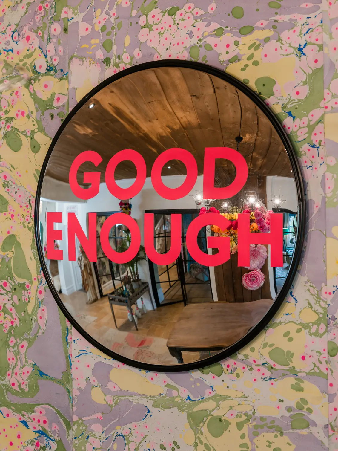 Round convex mirror with 'GOOD ENOUGH' hand painted in neon pink, against a pastel marbelled wallpapered wall