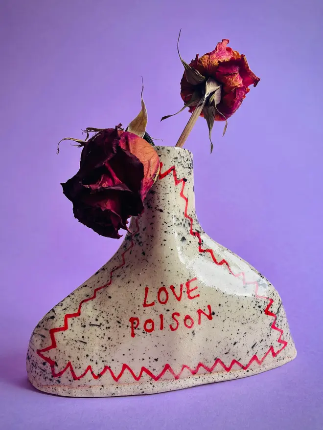 Love Potion/ Poison Bottle Vase, a vase that says 'love poison' in front of a purple backdrop. Each is stamped with a signature red dot on the bottom. 