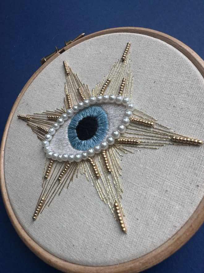 An angled embroidery hoop containing a stitched protective eye edged with pearl beads and a good sewn star with beads
