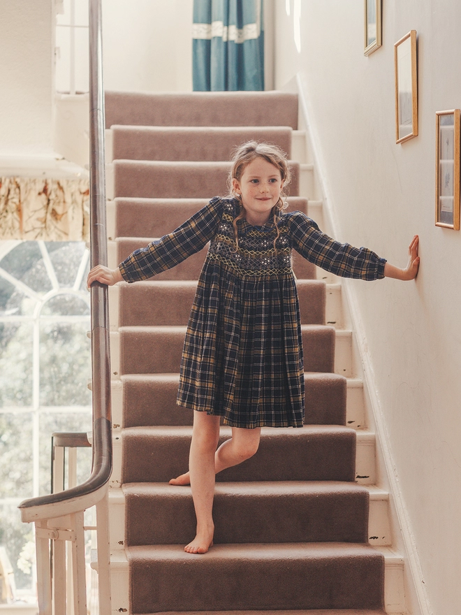 A girl stands on stairs wearing a navy and yellow dress with hand smocked detail