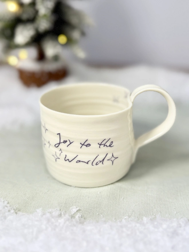 cream hand thrown porcelain mug with the words 'Joy to the World' printed onto the side with pretty line drawn stars. Sitting on a tabletop with snow, a mini lit Christmas tree 