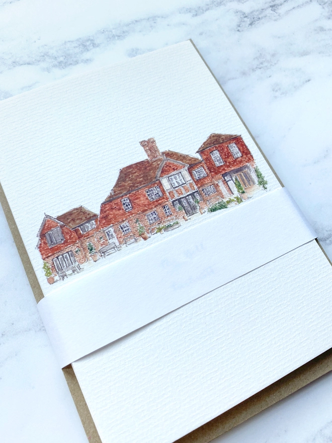 Greetings card showing watercolour painting of The Bell, in Ticehurst, a beautiful brick building with black and white framed window and door which is open welcoming you into the shop. The watercolour style is painted with a black pen outline and organic loose style with small details. The card sits on the top of a pack of cards with a white paper band around. 