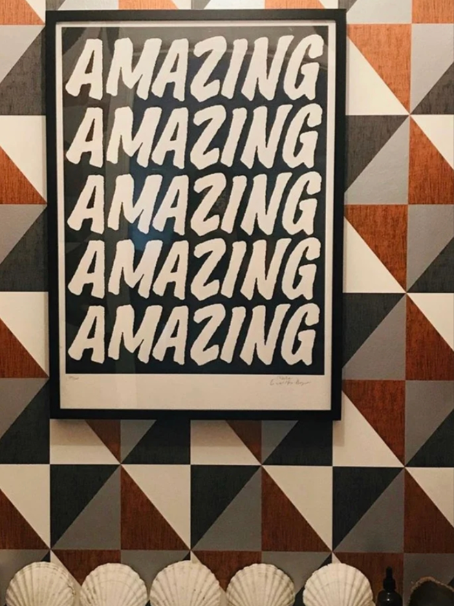 Black Amazing Poster. A framed print hanging against a tiled wall. The print has a black background and AMAZING written in a simple, all caps font five times.