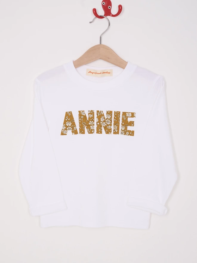 A white long sleeve cotton t-shirt appliquéd with the name Annie in a floral Liberty print, hanging on a hanger