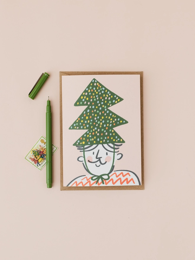 Christmas card featuring an illustration of a man wearing a Christmas tree hat on a pink background. 