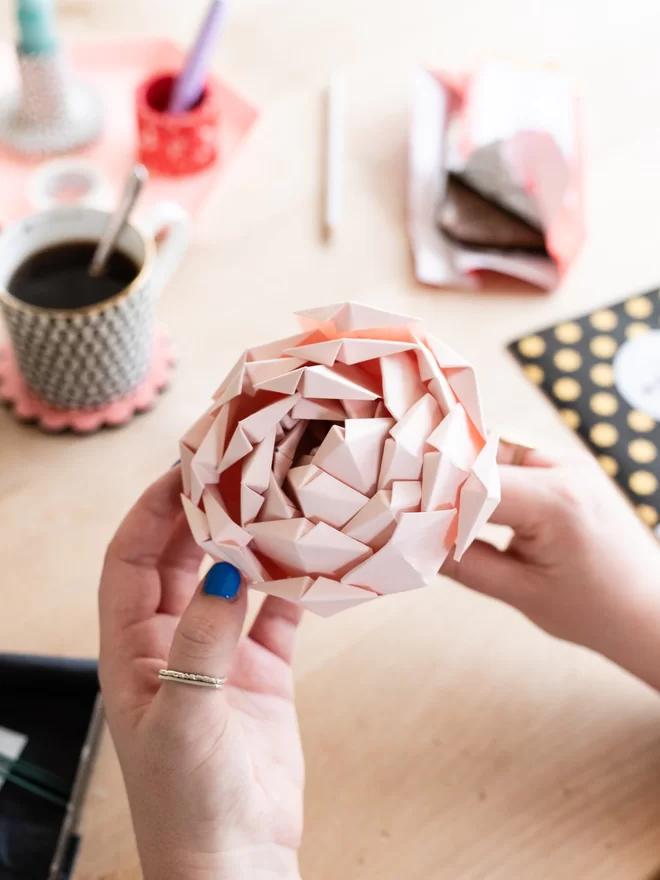 A blush origami peony flower held in hands of a maker, with a coffee cup, chocolate bar & stationery scattered on a desk.