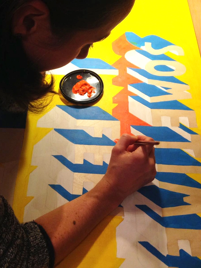 Artist Survival Techniques painting a partially finished typographic artwork that reads “SOMETIMES I FEEL TRULY AWFUL / FUCKING AMAZING” in uppercase letters. The artist is leaning forward, carefully applying orange paint to shadow of one of the bold 3D letters. The text features blue and white lettering with orange shadows on a yellow background.