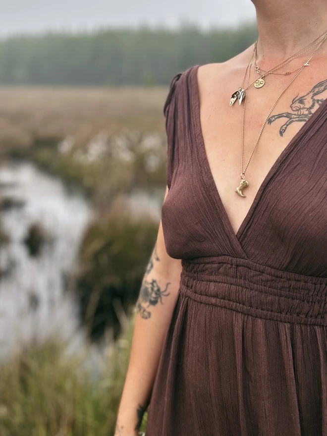 Image of a female torso wearing a brown dress and multiple necklaces, one necklace is a hand carved cowboy boot cast in gold toned brass. The background of the image is a bog like body of water.