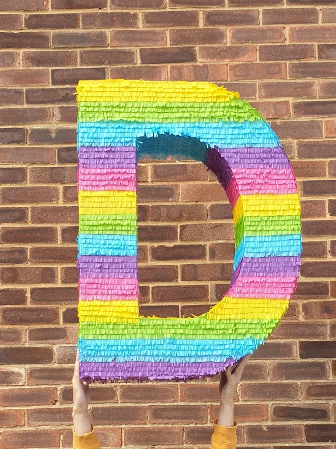 giant letter D pinata in rainbow stripes held up in front of a brick wall