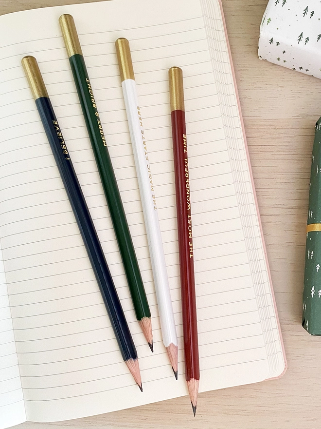 Four festive pencils lay on an open notebook on a wooden desk.