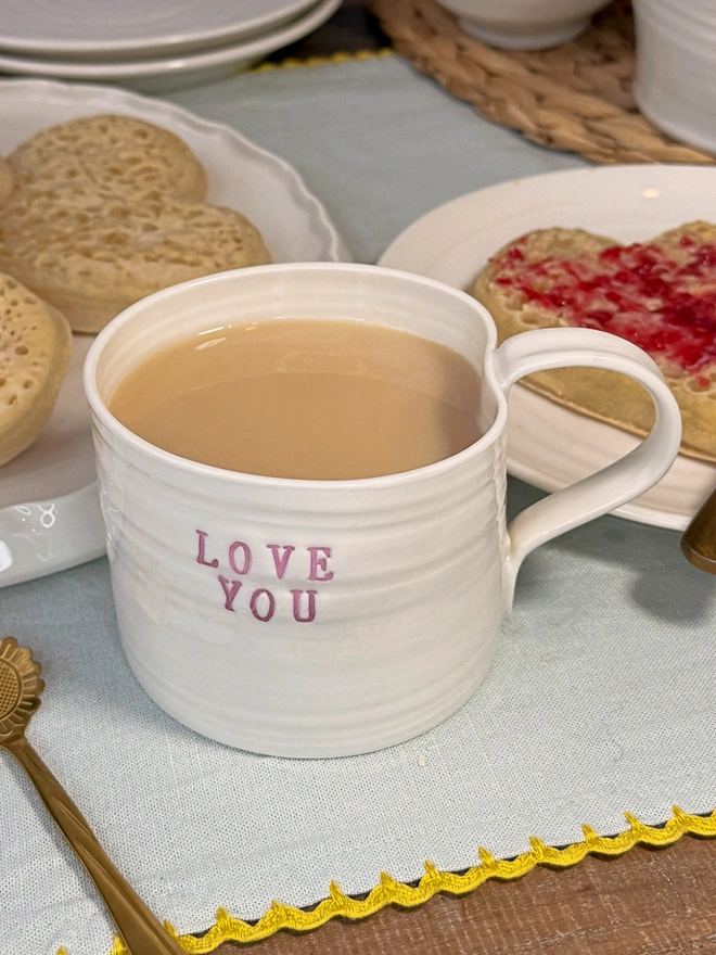 hand thrown porcelain cream mug with pink lustre text on the side that says LOVE YOU. The mug is sat upon a sage table cloth amongst a tea time scene of heart shaped crumpets
