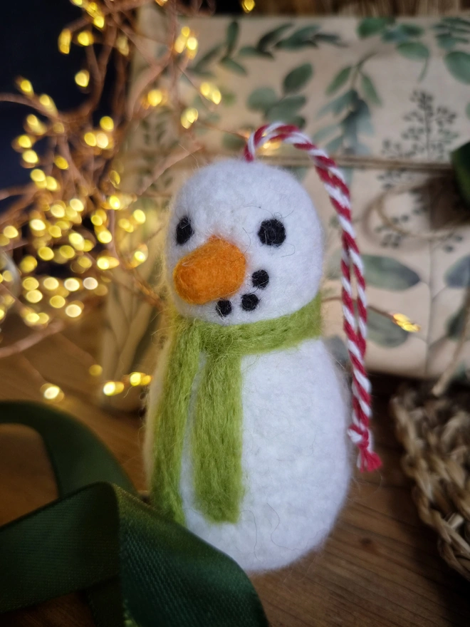 A hanging Christmas decoration ornament in the shape of a white felt snowman with a green scarf.
