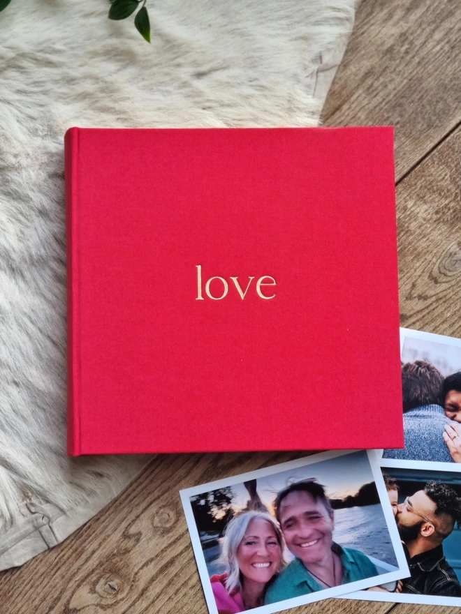 A hand-bound red photo album printed with the word 'love' in gold lettering rests on a fur rug and a wooden floor. It is surrounded by family photographs.