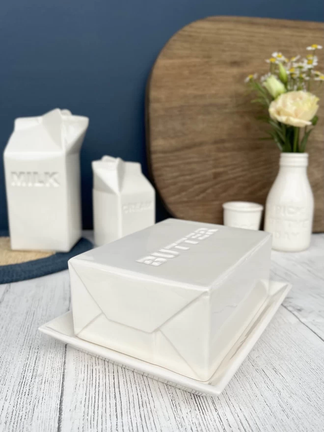 A handmade ceramic butter dish lid, emulates a pat butter in it packaging sits on an oblong base.